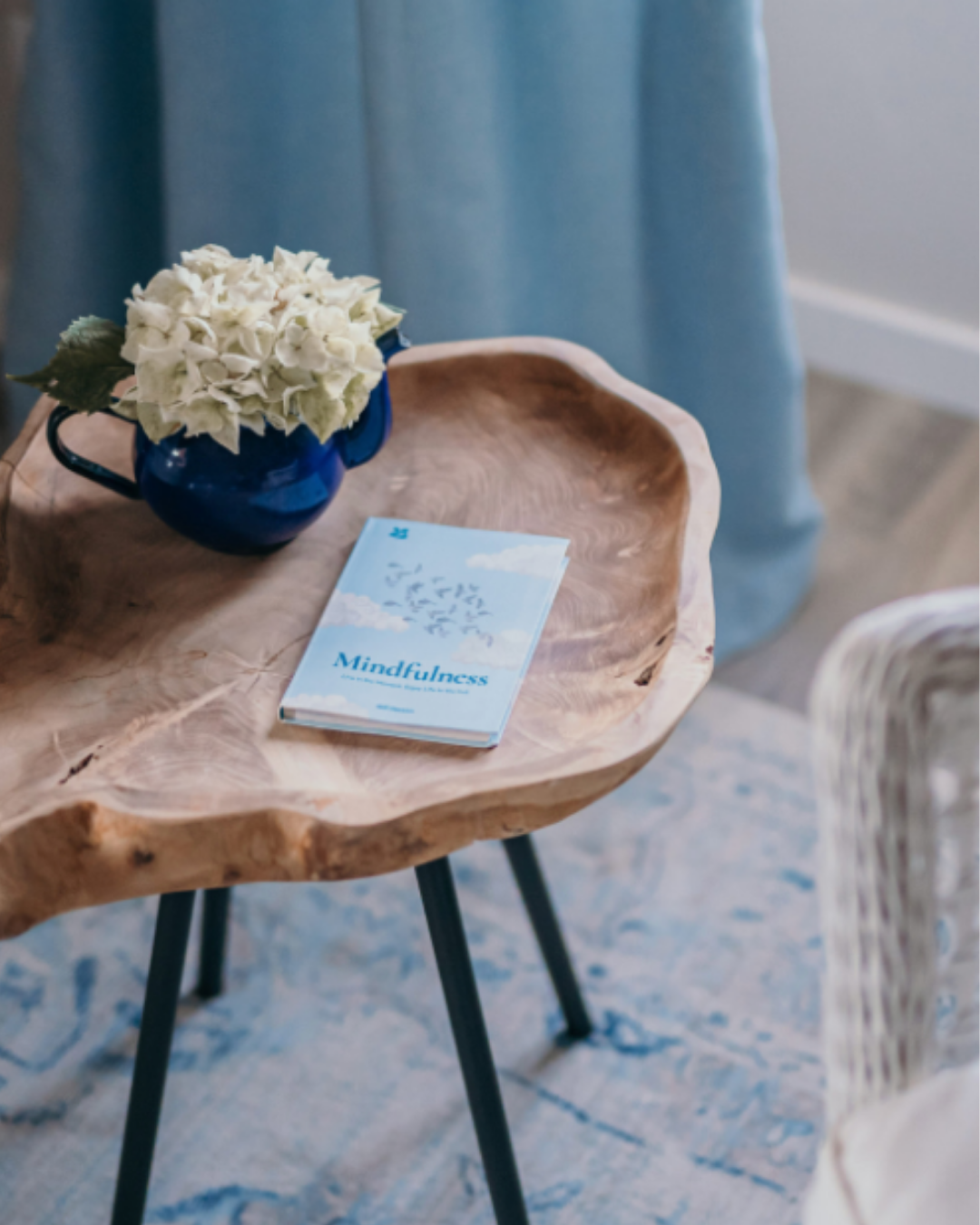 Close-up of a wooden side table with a blue ceramic jug holding white flowers and a light blue book titled “Mindfulness” resting on top, set against a blue curtain backdrop.