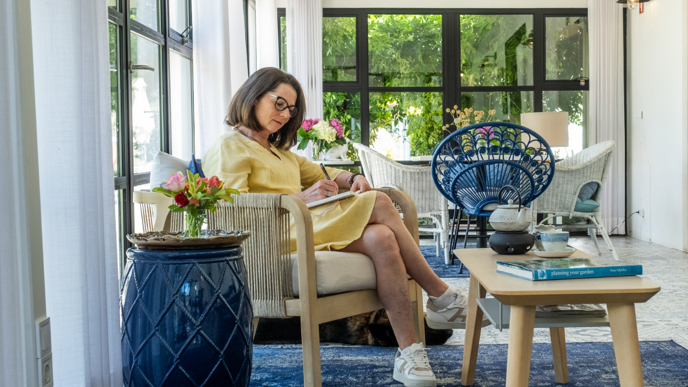 Woman in a yellow dress sitting in a bright, airy living room, writing in a notebook with a pen, surrounded by flowers, wicker furniture, and natural light streaming through large windows.