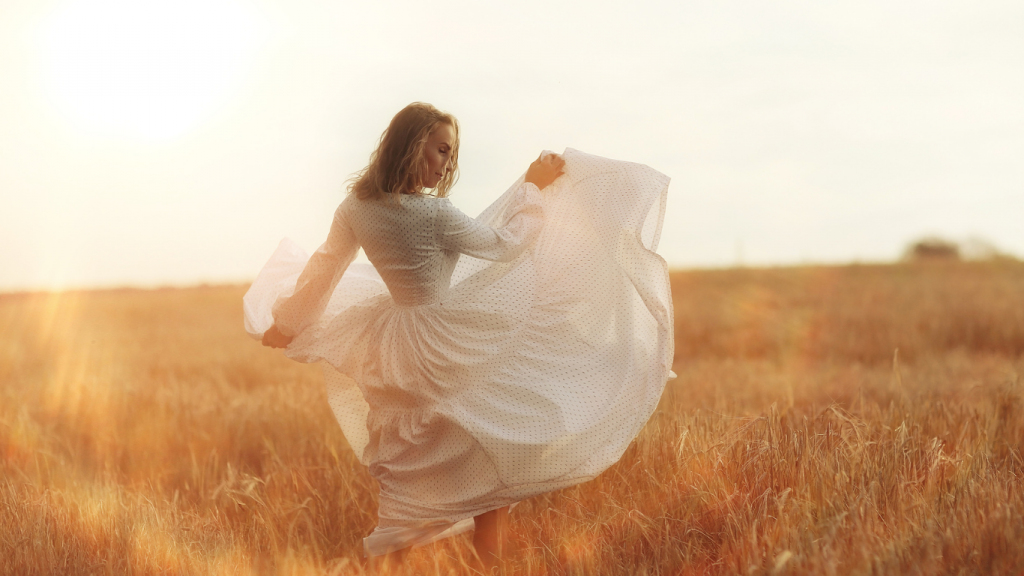 Woman in a flowing white dress twirling gracefully in a golden wheat field at sunset, with warm sunlight creating a soft, dreamy glow.